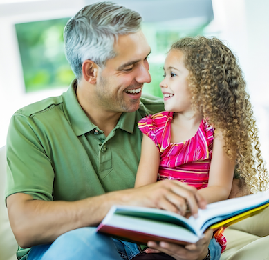 Father reading a book to his daughter as she sits on his lap. They are smiling at each other